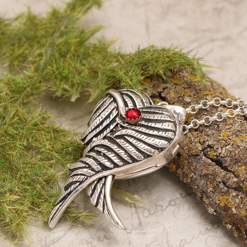 Pu&ograve; includere: Collana con ciondolo in argento a forma di cuore con ali dettagliate e un accento di pietra preziosa rossa.