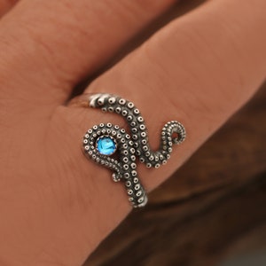 May include: A silver ring shaped like an octopus tentacle that wraps around the finger. The tentacle features textured suction cups and is accented with a single, small, round, bright blue gemstone set within the coiled section of the metal.