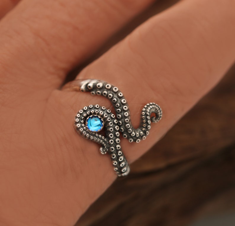 May include: A silver ring shaped like an octopus tentacle that wraps around the finger. The tentacle features textured suction cups and is accented with a single, small, round, bright blue gemstone set within the coiled section of the metal.