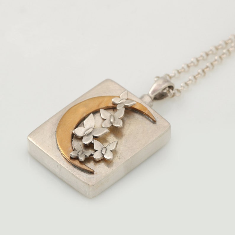 Op de afbeelding: Een zilveren ketting met een rechthoekige hanger met een halve maan en gestileerde vlinders. De maan is van goud.