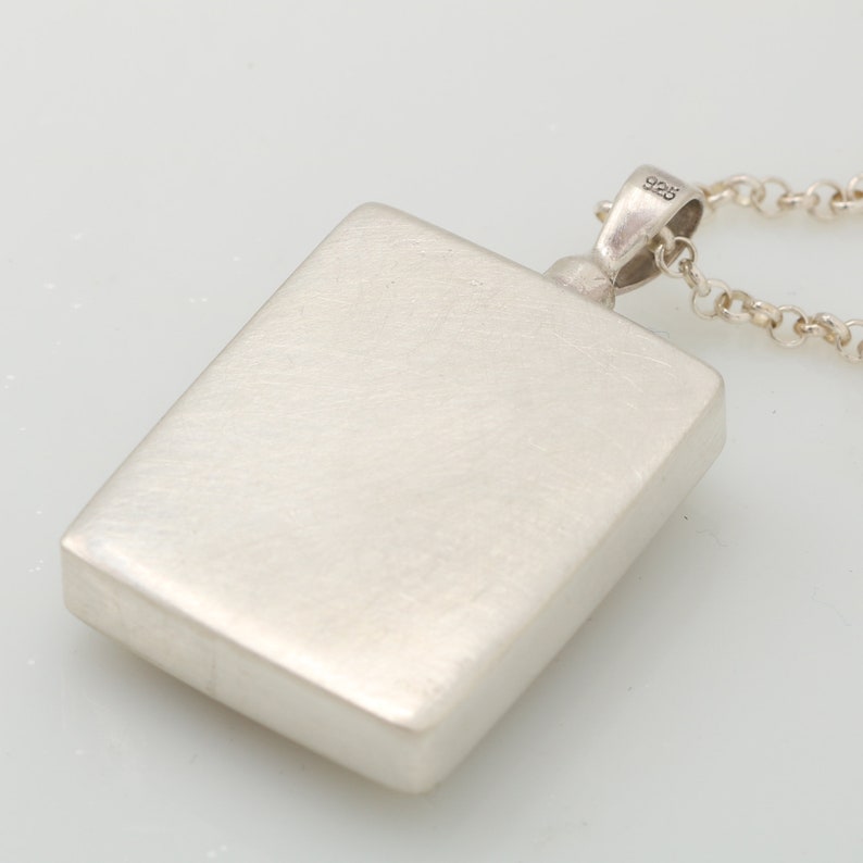 Op de afbeelding: Een zilveren hanger met een vierkante vorm en een gladde, geborstelde afwerking. De hanger is bevestigd aan een zilveren ketting met een karabijnhaak. De hanger is aan de achterkant gemarkeerd met het nummer "925".