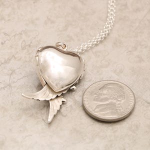 Pu&ograve; includere: Collana con ciondolo in argento con un ciondolo a forma di cuore e ali d'angelo. Il ciondolo &egrave; aperto e il retro &egrave; visibile. La collana &egrave; accanto a una moneta da 25 centesimi di dollaro americano per il confronto delle dimensioni.