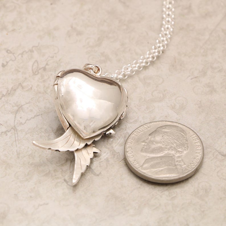 Pu&ograve; includere: Collana con ciondolo in argento con un ciondolo a forma di cuore e ali d'angelo. Il ciondolo &egrave; aperto e il retro &egrave; visibile. La collana &egrave; accanto a una moneta da 25 centesimi di dollaro americano per il confronto delle dimensioni.