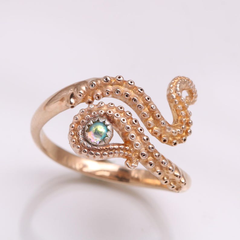 May include: A rose gold-toned ring featuring a textured octopus tentacle design that wraps around the finger. The tentacle surface is covered in small, raised suction cups, and a single, small, iridescent round stone is set within the coil.