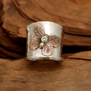K&ouml;nnte beinhalten: Ein silberner Ring mit geh&auml;mmerter Textur zeigt einen kupfernen Schmetterling mit einem blauen Edelstein in der Mitte.