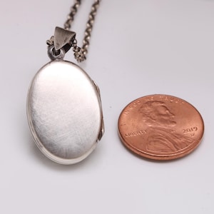 May include: A silver-toned oval locket pendant suspended from a matching chain, featuring a brushed metallic finish. The locket is positioned next to a copper-colored United States one-cent coin for scale, providing a clear visual size reference.