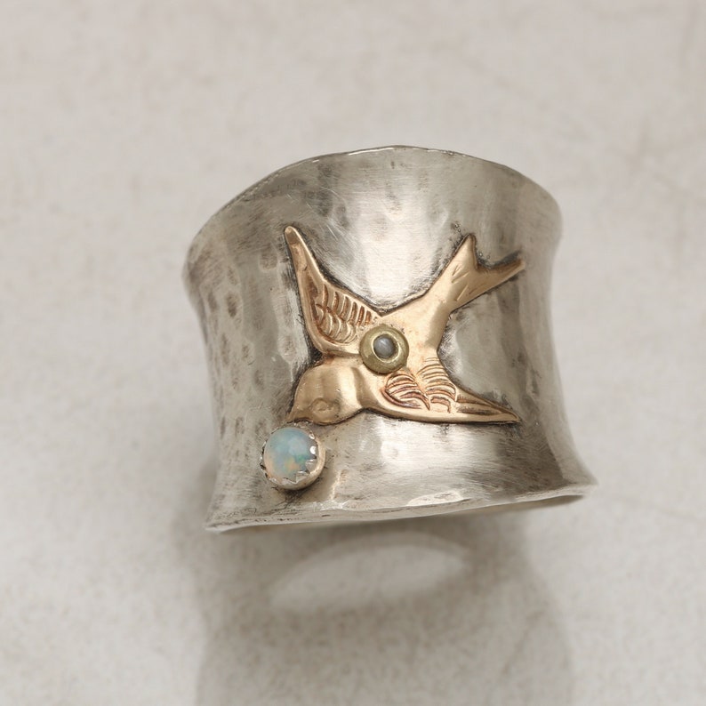 May include: A silver ring with a hammered finish features a gold bird with an opal gemstone. The bird is in flight and has a detailed design.