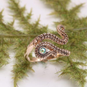 May include: A copper-toned metal ring shaped like an octopus tentacle that wraps around the finger. The surface features a textured pattern of small circular suckers and is accented with a single, small, iridescent, round, multi-colored gemstone.