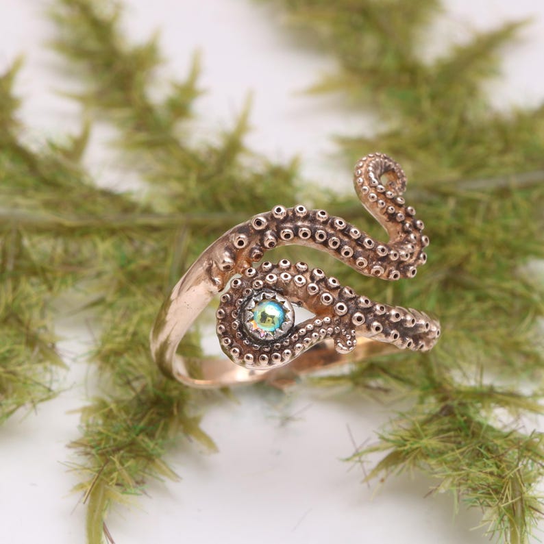 May include: A copper-toned metal ring shaped like an octopus tentacle that wraps around the finger. The surface features a textured pattern of small circular suckers and is accented with a single, small, iridescent, round, multi-colored gemstone.