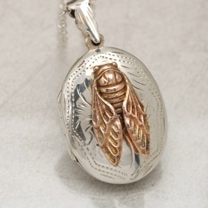 May include: Silver locket necklace with a detailed floral design and a gold-toned cicada charm.
