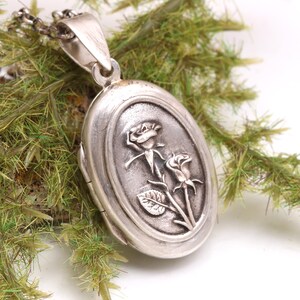 May include: An oval silver locket pendant featuring a raised relief design of two roses on the front surface. The locket has a polished metallic finish with darkened recessed areas to highlight the floral details and includes a silver chain bail.
