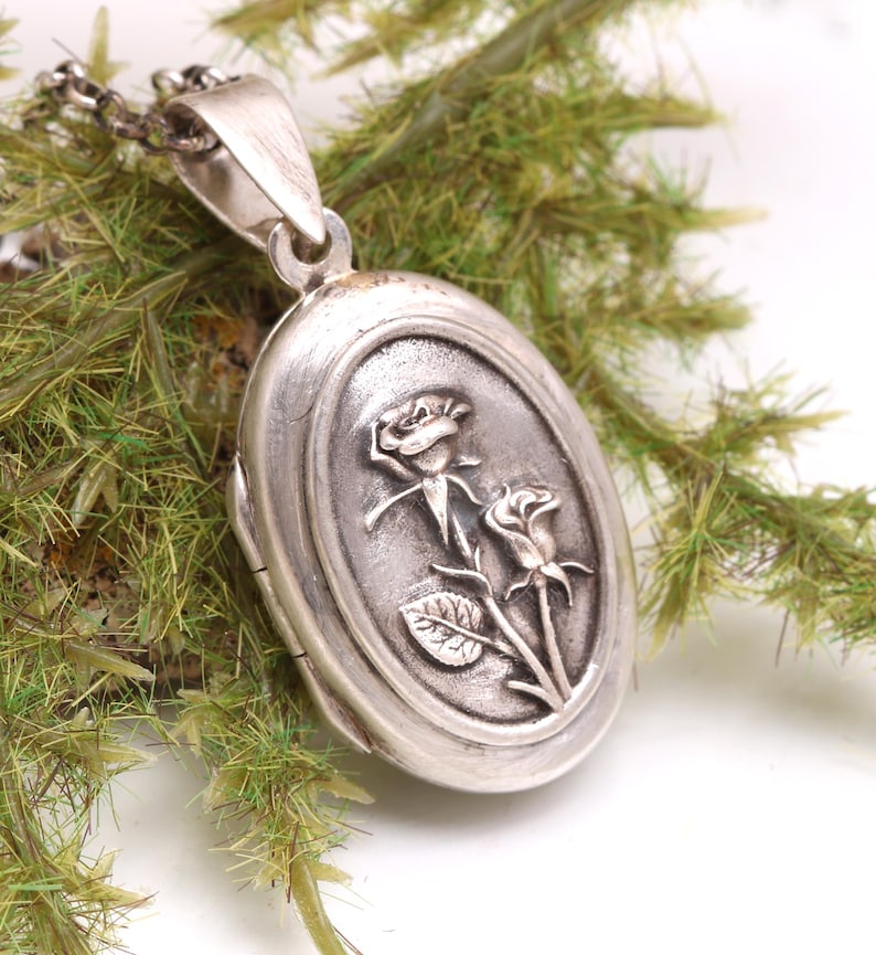 May include: An oval silver locket pendant featuring a raised relief design of two roses on the front surface. The locket has a polished metallic finish with darkened recessed areas to highlight the floral details and includes a silver chain bail.
