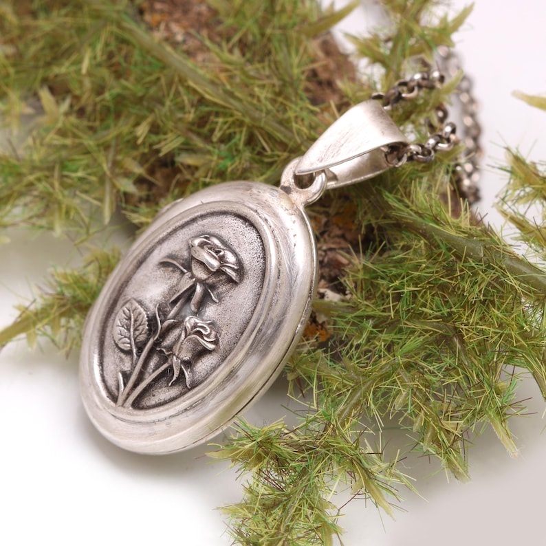May include: An oval silver locket pendant featuring a raised relief design of two roses with stems and leaves. The locket has a polished silver finish with darkened recessed areas to highlight the floral detail and is attached to a silver chain.