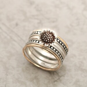 May include: A silver ring featuring a sunflower design. The ring is made up of three bands, one of which is gold. The sunflower is brown and has a textured centre.