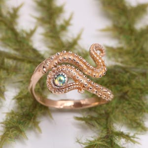 May include: A rose gold-toned ring featuring a textured octopus tentacle design that wraps around the finger. The band includes a small, round, iridescent gemstone set within the tentacle structure, showcasing detailed suction cup patterns.