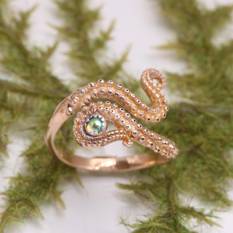 May include: A rose gold-toned ring featuring a textured octopus tentacle design that wraps around the finger. The band includes a small, round, iridescent gemstone set within the tentacle structure, showcasing detailed suction cup patterns.