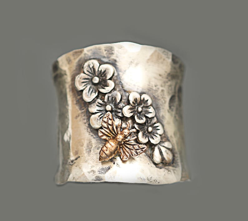 Puede incluir: Un anillo de plata con una superficie texturizada y un dise&ntilde;o de flores y una abeja. La abeja es de oro.