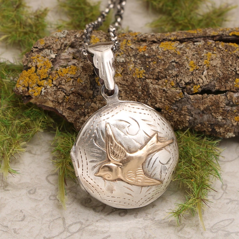 May include: A silver locket necklace with a detailed design and a gold bird charm. The locket is round and has a hinged lid. The bird is in flight and has a detailed design.