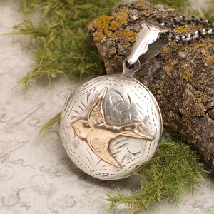 May include: Silver locket necklace with a gold bird design. The locket is engraved with a floral pattern and has a hinged lid.