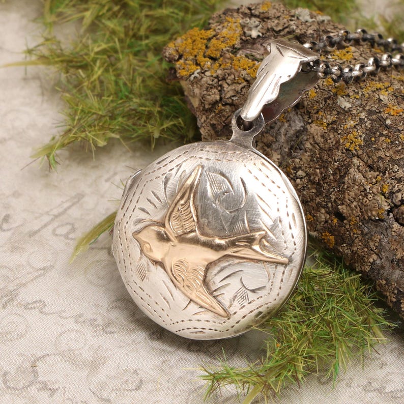 May include: Silver locket necklace with a gold bird design. The locket is engraved with a floral pattern and has a hinged lid.
