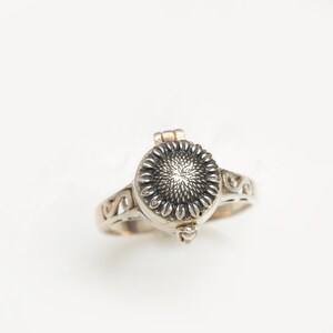 May include: A silver ring featuring a sunflower design. The ring has a hinged lid that opens to reveal a small compartment.
