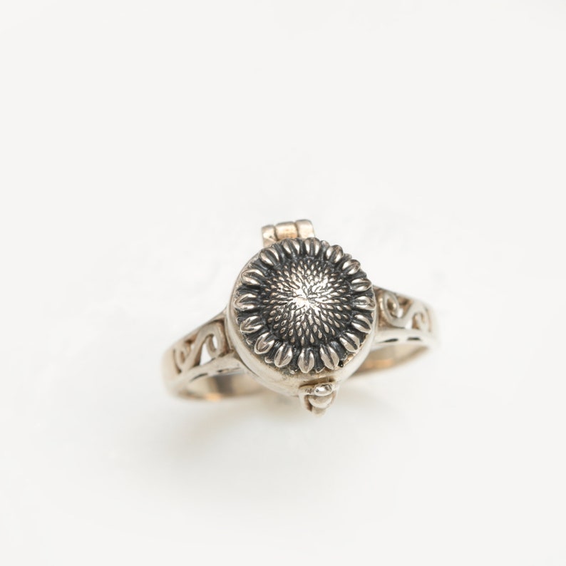 May include: A silver ring featuring a sunflower design. The ring has a hinged lid that opens to reveal a small compartment.