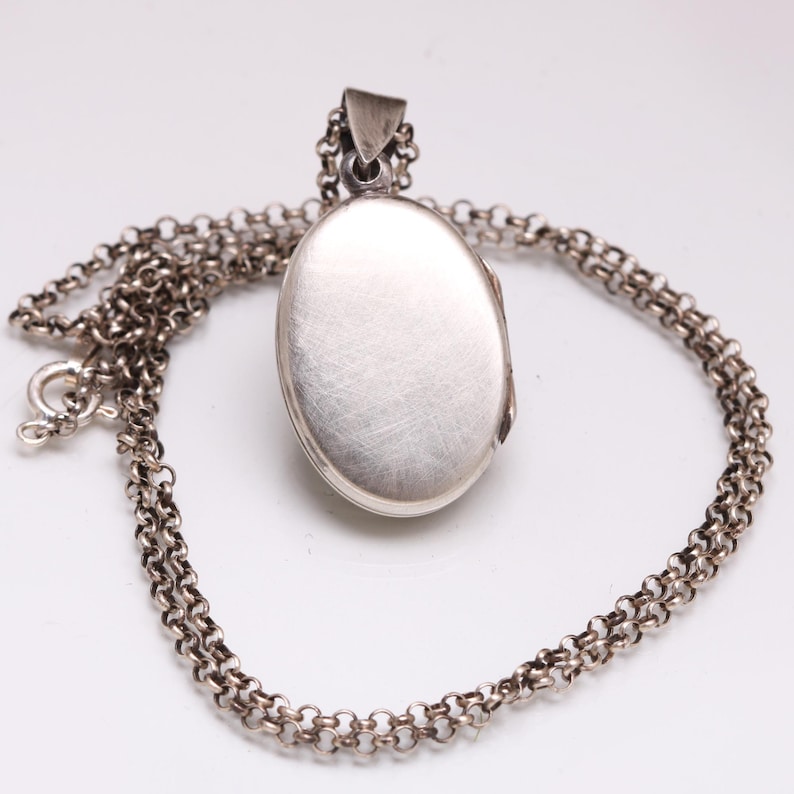 May include: An oval-shaped silver locket pendant with a brushed metallic finish, suspended from a matching silver rolo chain. The locket features a simple, smooth surface and a secure bail at the top, connected to a circular spring ring clasp.