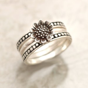 May include: Silver ring with a three-band design. The top band features a raised sunflower design. The other two bands have a dotted texture.