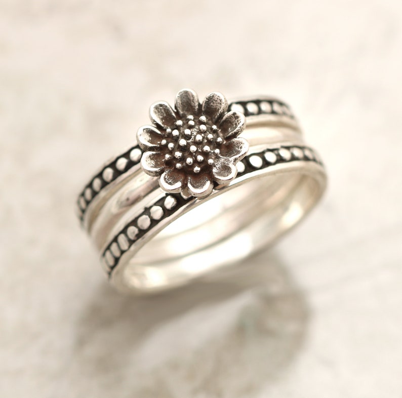 May include: Silver ring with a three-band design. The top band features a raised sunflower design. The other two bands have a dotted texture.