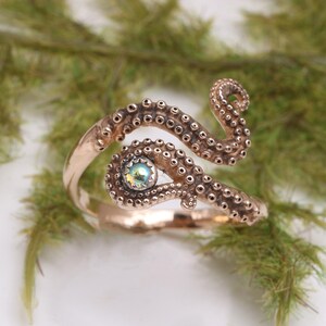 May include: A metallic copper-toned ring shaped like an octopus tentacle that wraps around the finger. The design features raised circular suction cups along the length of the band and a single iridescent, round stone set in the center.