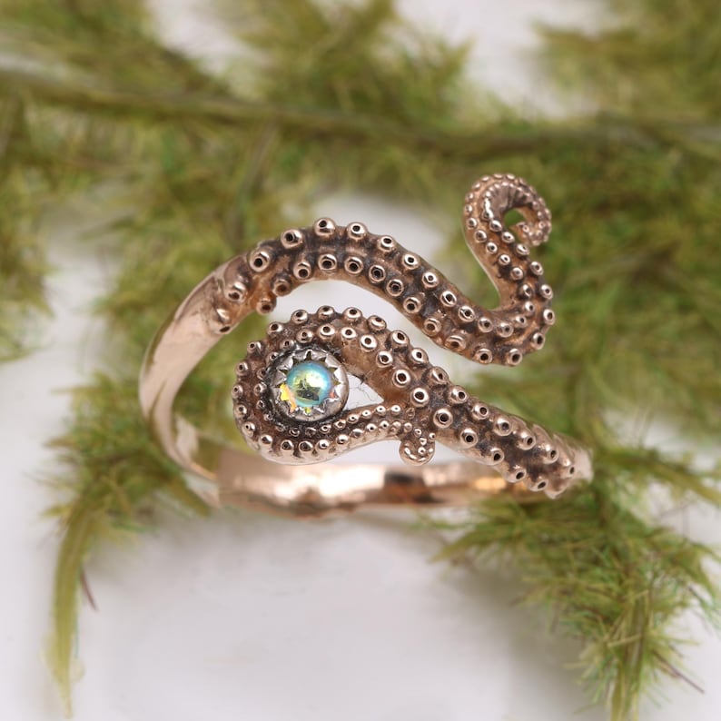 May include: A metallic copper-toned ring shaped like an octopus tentacle that wraps around the finger. The design features raised circular suction cups along the length of the band and a single iridescent, round stone set in the center.