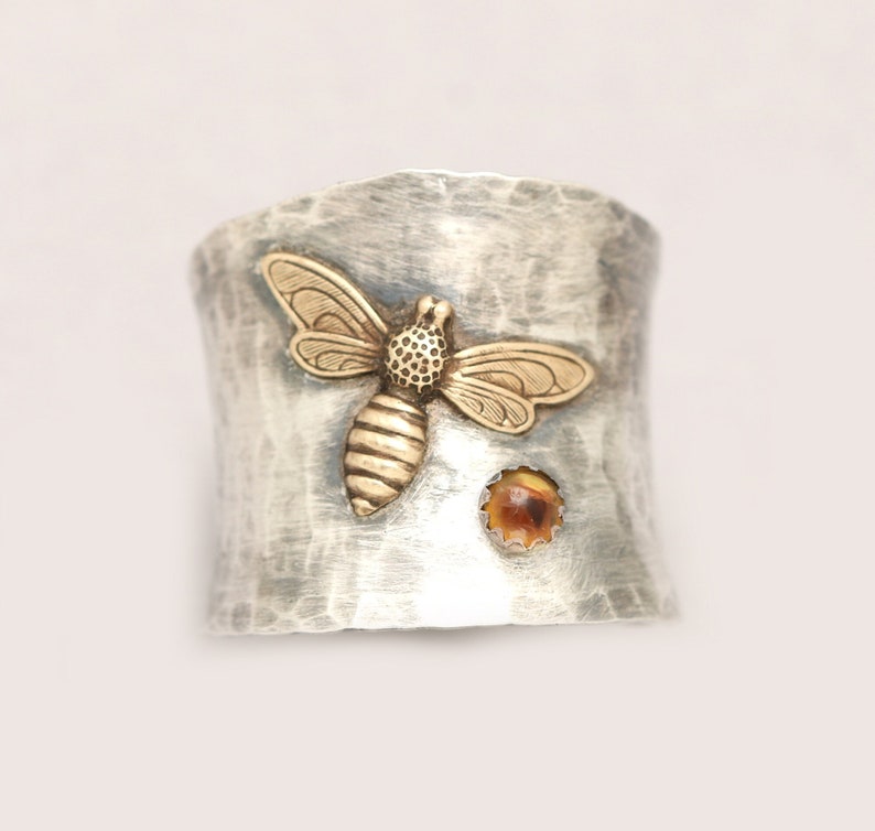 Puede incluir: Un anillo de plata con textura martillada presenta una abeja de oro y una peque&ntilde;a piedra &aacute;mbar.