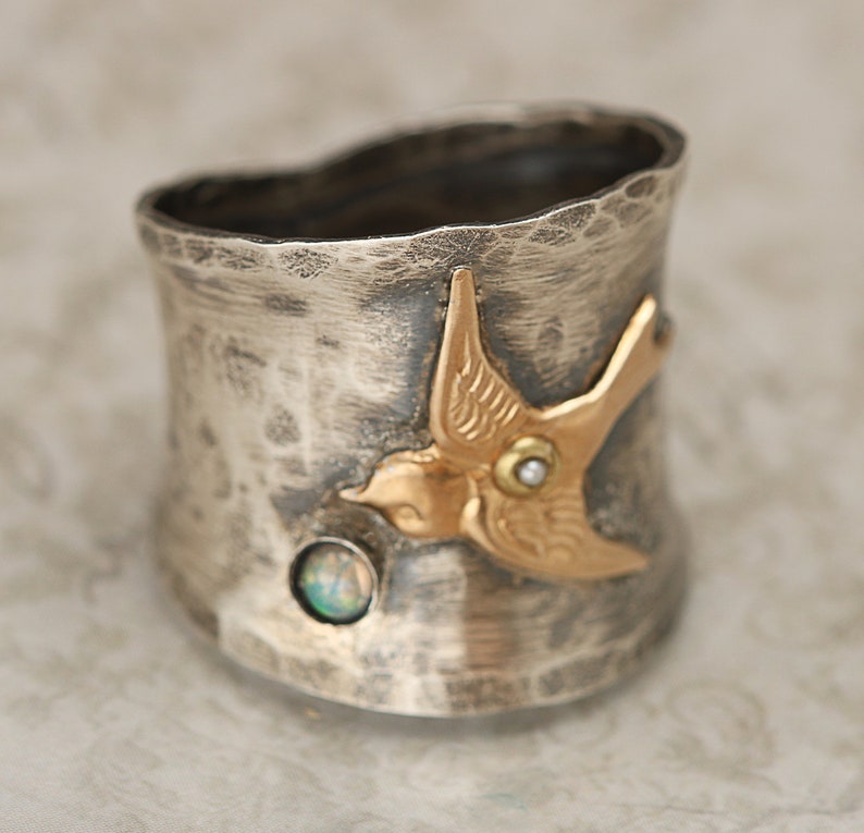 May include: A silver ring with a hammered finish features a gold bird with a pearl and opal accents.