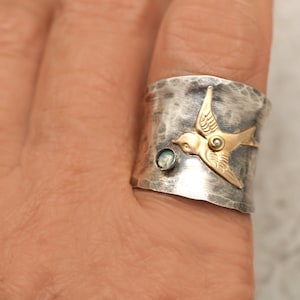 Puede incluir: Un anillo de plata con acabado martillado presenta un p&aacute;jaro de oro con una piedra preciosa de &oacute;palo. El anillo se lleva en un dedo.