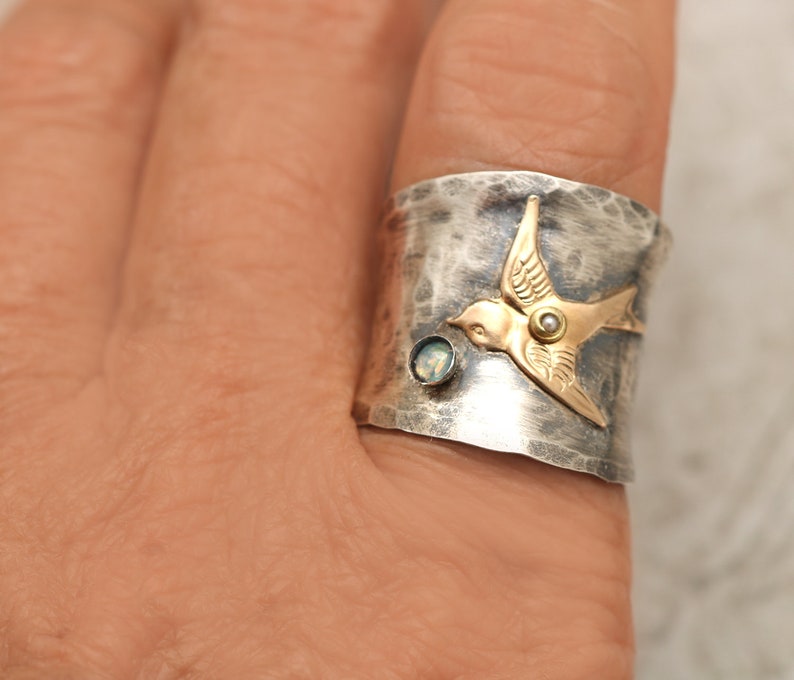 Puede incluir: Un anillo de plata con acabado martillado presenta un p&aacute;jaro de oro con una piedra preciosa de &oacute;palo. El anillo se lleva en un dedo.