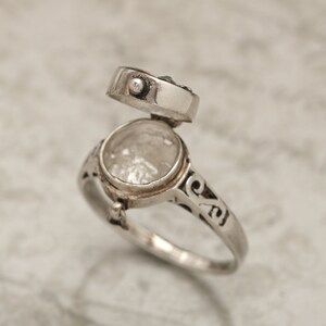 May include: A silver ring with an open hinged compartment. The ring has an intricate design and is set with a small blue stone.