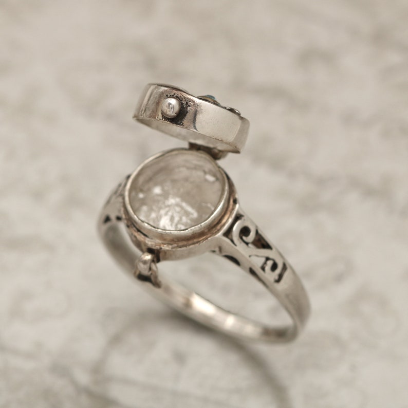 May include: A silver ring with an open hinged compartment. The ring has an intricate design and is set with a small blue stone.