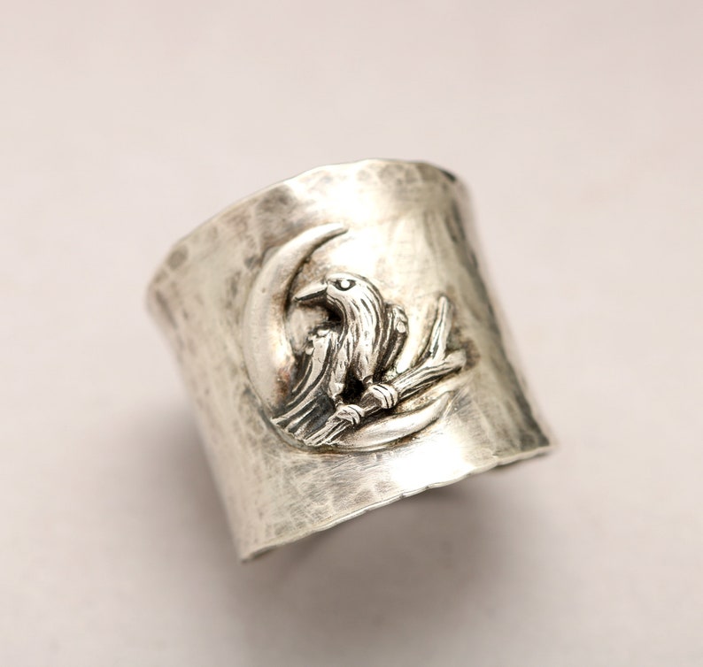 K&ouml;nnte beinhalten: Ein silberner Ring mit geh&auml;mmerter Textur zeigt ein erhabenes Design eines Vogels, der auf einem Ast sitzt, mit einem Halbmond dahinter.