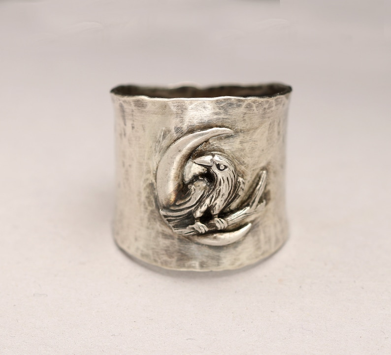 K&ouml;nnte beinhalten: Ein silberner Ring mit einer strukturierten Oberfl&auml;che und einem Design eines Rabens, der auf einem Halbmond sitzt.