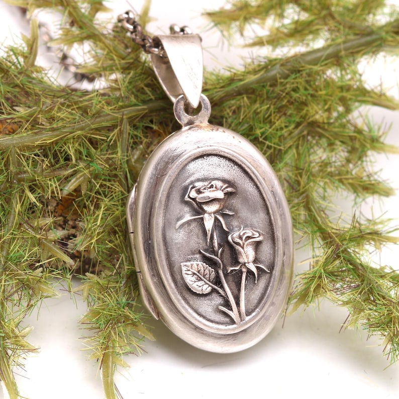May include: An oval-shaped silver locket pendant featuring a raised relief design of two roses on the front surface. The locket is attached to a silver chain and has a polished metallic finish with darkened recessed areas to highlight the floral detail.