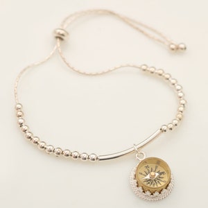 May include: A silver beaded bracelet with a compass charm. The bracelet has a sliding knot closure and is adjustable in size.