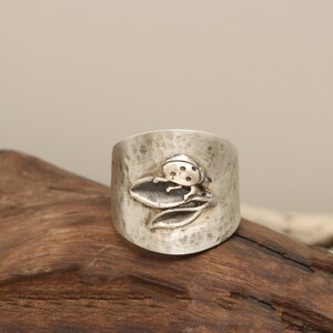 May include: A silver ring with a hammered finish featuring a ladybug design. The ladybug is depicted on a leaf with a textured surface.