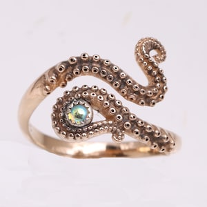 May include: A gold-toned metal ring shaped like an octopus tentacle that wraps around the finger. The tentacle features detailed suction cups along its length and holds a single, small, iridescent, round gemstone set in a silver-colored bezel.