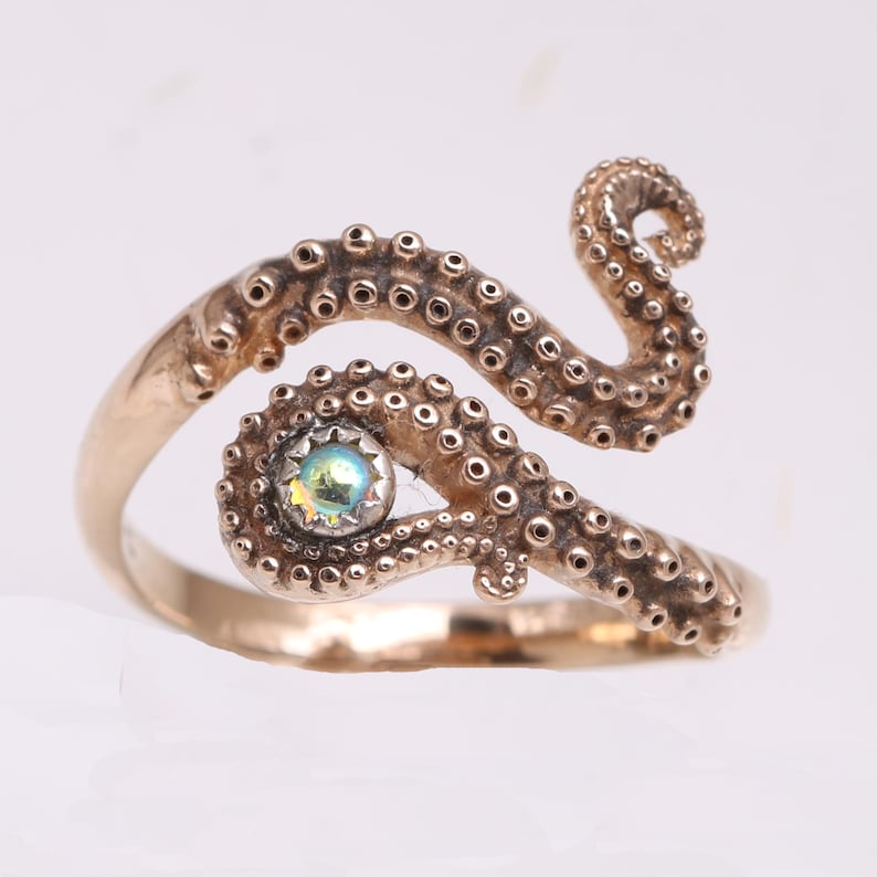 May include: A gold-toned metal ring shaped like an octopus tentacle that wraps around the finger. The tentacle features detailed suction cups along its length and holds a single, small, iridescent, round gemstone set in a silver-colored bezel.