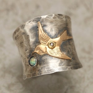 May include: A silver ring with a hammered finish features a gold bird with a pearl accent. The bird is set against a green opal stone.