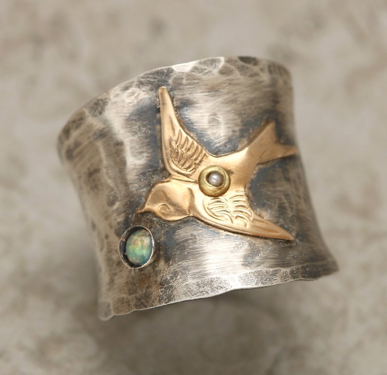 May include: A silver ring with a hammered finish features a gold bird with a pearl accent. The bird is set against a green opal stone.
