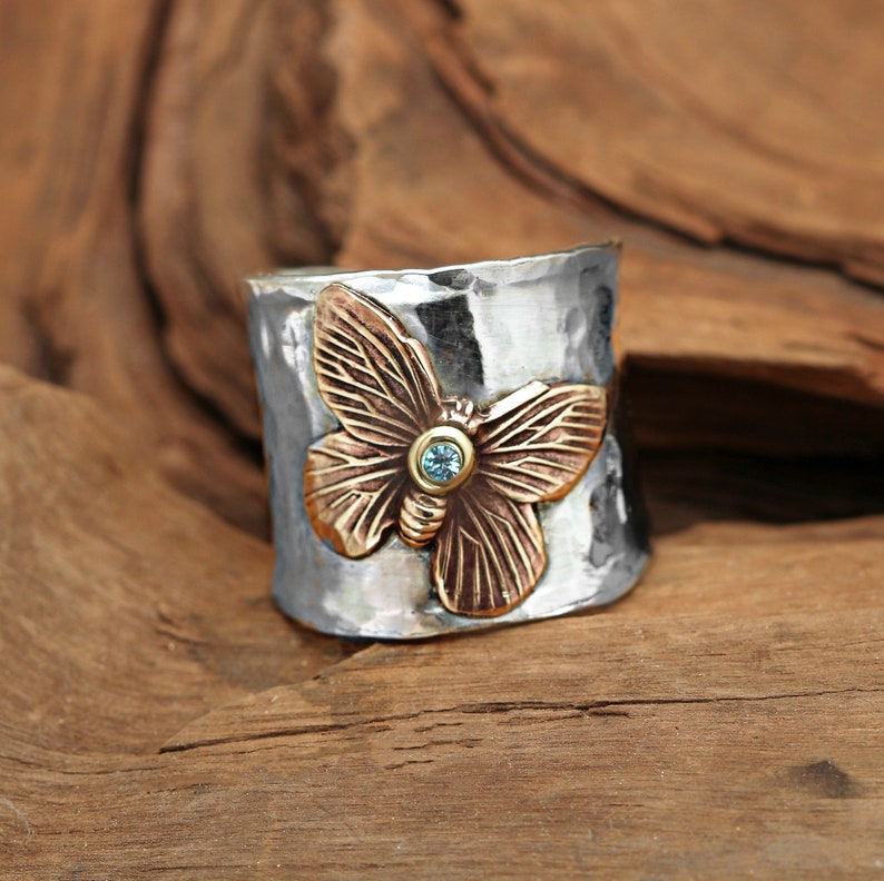 K&ouml;nnte beinhalten: Ein silberner Ring mit geh&auml;mmerter Oberfl&auml;che zeigt einen bronzenen Schmetterling mit einem blauen Edelstein in der Mitte.