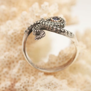 May include: A silver ring shaped like an octopus tentacle that wraps around the finger. The metal has a dark, oxidized finish highlighting the textured suckers along the tentacle. A small, round, dark blue gemstone is set into the top section.