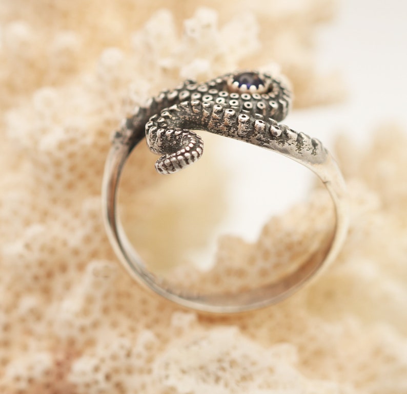 May include: A silver ring shaped like an octopus tentacle that wraps around the finger. The metal has a dark, oxidized finish highlighting the textured suckers along the tentacle. A small, round, dark blue gemstone is set into the top section.