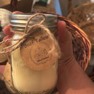 Op de afbeelding: Een heldere glazen pot gevuld met een romige, gebroken witte substantie, waarschijnlijk talg. De pot heeft een zilveren deksel en een bruin papieren label met de tekst "Beef Tallow", vastgebonden met touw.